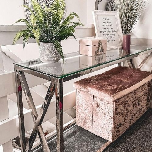 glass and chrome console table with X design to either end of leg frame, in a bright landing, with pot plant, photo and keepsake box, and pink velvet storage box stool placed underneath. 