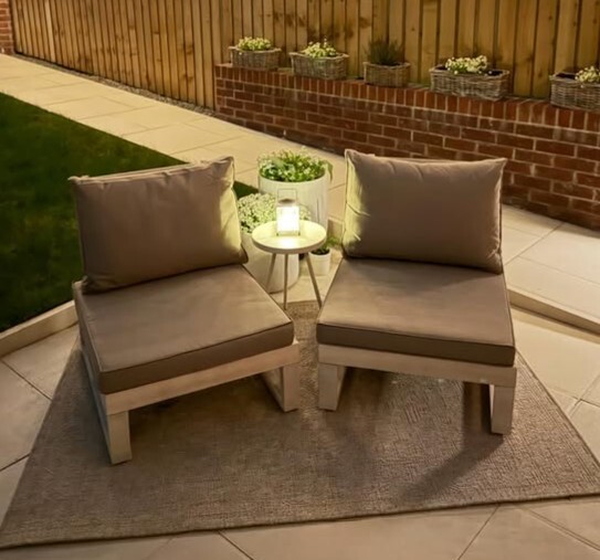 2 simple whitewash and grey wood garden chairs and side table in evening garden with lamp on table and planters on wall behind