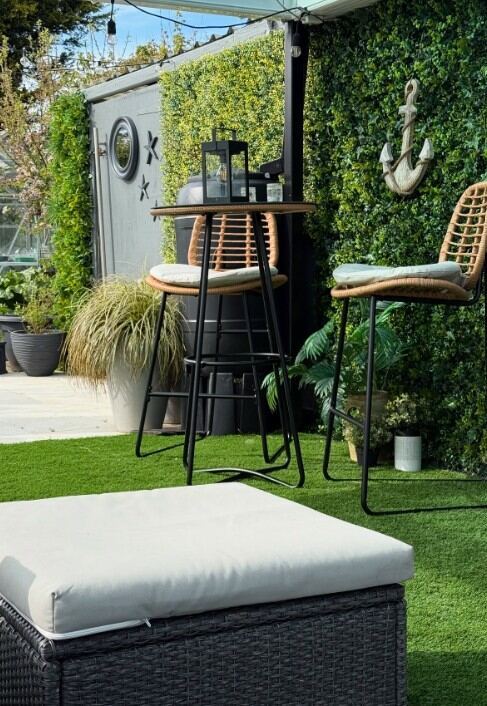 2 seat round rattan bar table and chairs set in modern garden against leafy wall with anchor decor and lantern on table