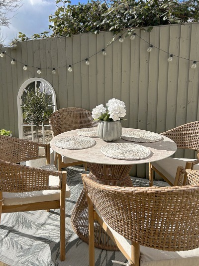 Birdgerton inspired garden dining set with round marble and rattan garden table, matching dining chairs and white roses in vase on table. Arch window garden mirror behind with string lights