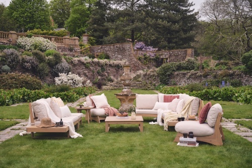 wooden garden corner sofa and accent chairs with plump cream cushions, dressed with ducky pink and terracotta throws and scatter cushions, on a green lawn with stone wall behind