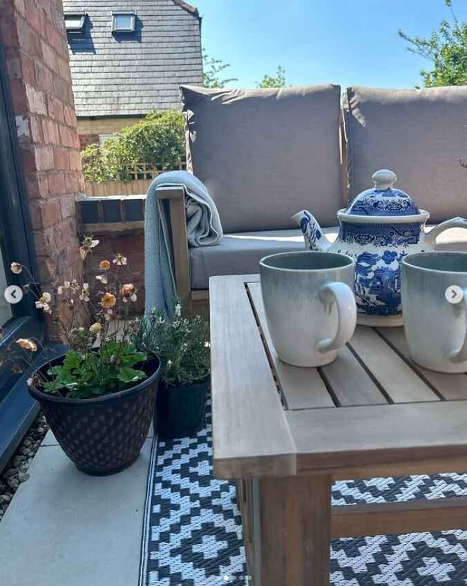wooden garden sofa and table set on balcony, with tea set on table, garden rug beneath table, plant pots and brick wall