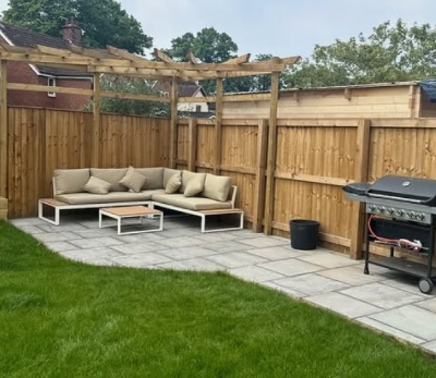 white metal corner gardne sofa with beige cushions beneath pergods, next to BBQ on garden lawn and patio area