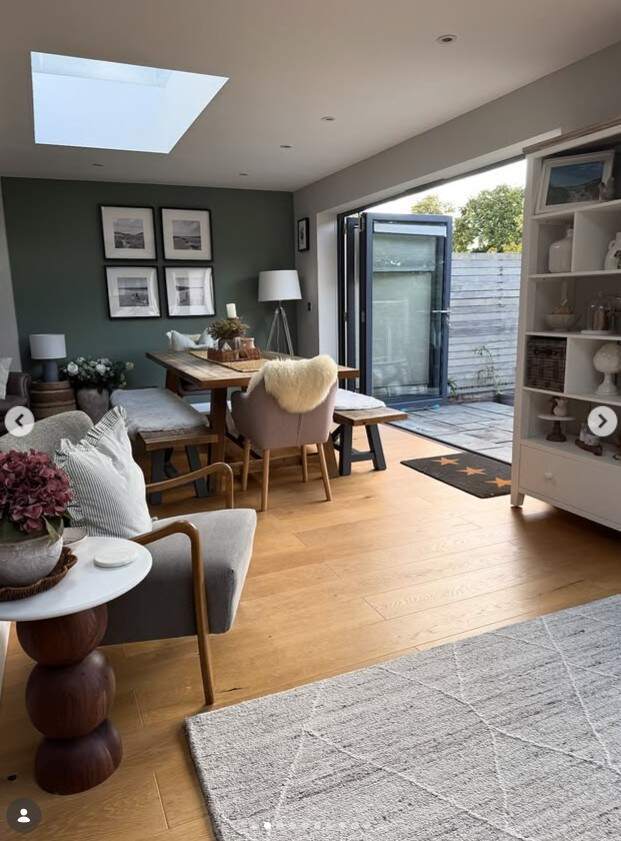 modern cottagecore style living room and dining space with accent chair, round side table and wood dining table with benches and chairs in background.Green and cream walls, wood floor, grey rug.