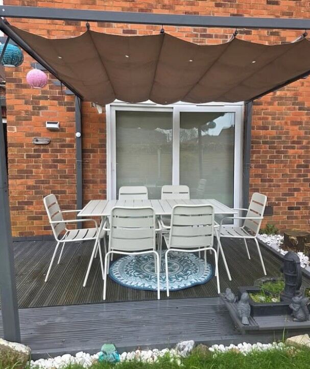 cream metal slatted garden dining table with 6 matching metal garden dining chairs on wood deck, with round rug beneath, with pergoda awning overhead, brick wall and French windows behind.