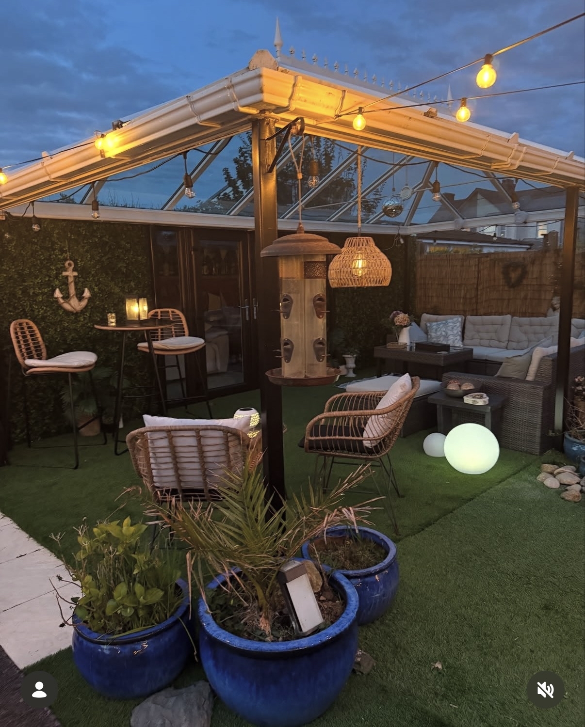 garden at dusk with bar table & chair, and garden sofa under pergoda, with string lights, lanterns and garden lighting set up