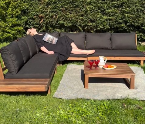 8 seat wood garden corner sofa in walnut wood with black cushions on green lawn with woman laying on sofa with a magazine