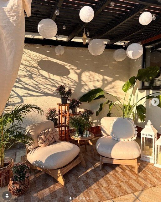 2 wood garden lounge accent chairs with thick cream cushions in covered outdoor patio area, with lights and lanterns and potted plants