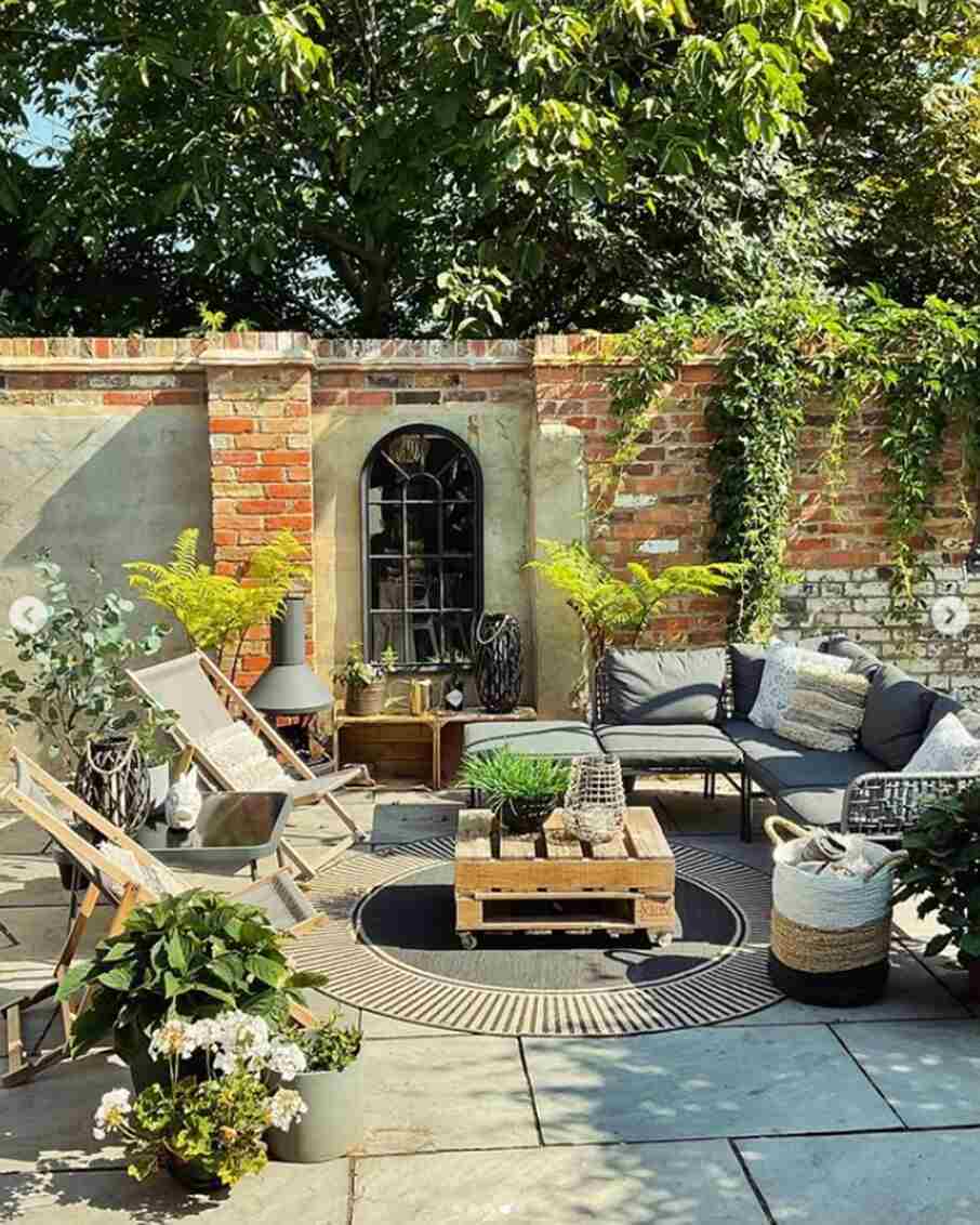 pretty brick walled garden with grey rattan garden sofa, deckchairs and wooden pallet coffee table, outdoor mirror and lots of potted plants