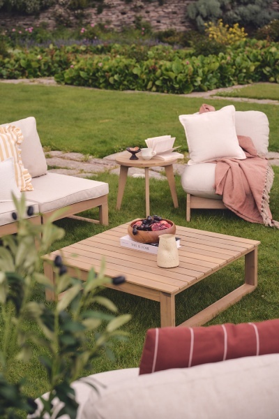garden seating ideas with wood garden seats, cream cushions and wooden garden coffee table with terracotta and pink throws and scatter cushions on lawn
