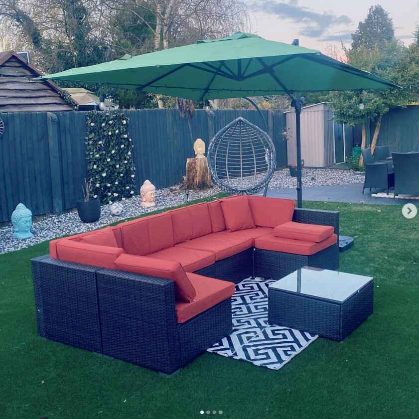 brown rattan modular garden sofa with orange cushions, with parasol and garden rug in forground on grass. Egg chair and garden dining set visible in background. 