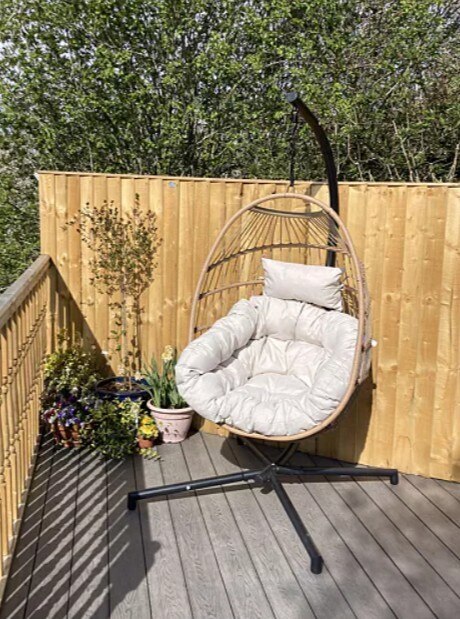 Beige Rattan & Cream Garden Hanging Egg Chair Seat on wood patio deck with planting and fence behind