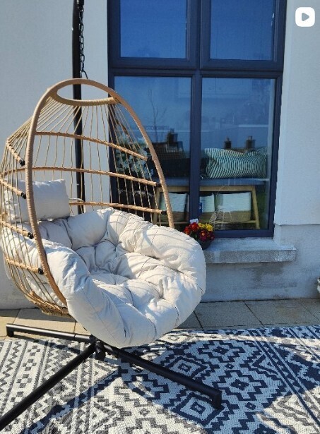 beige rattan hanging egg chair on black metal frame, on outdoor rug beside a wall