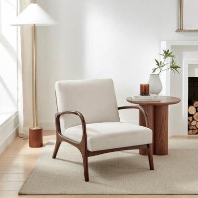 mid-century modern accent chair in cream fabric with wood arms, beside round wood side table and with wood and brass standing floor lamp behind