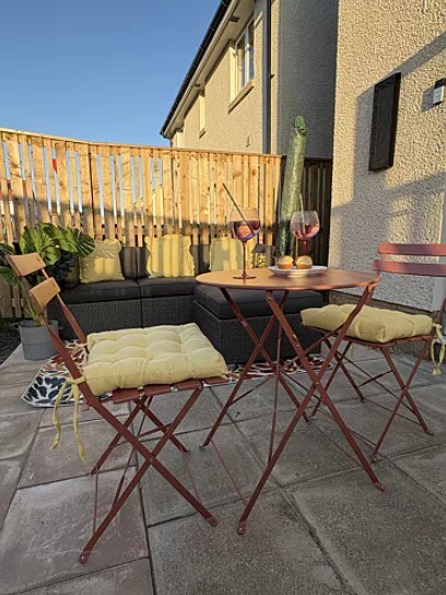 Red Metal 2 Seat Garden Bistro Table Set with yellow cushions on paved garden patio with fence and fairy lights behind