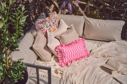 metal sunlounger day bed with cosy neutral pillows and throws, and 2 colourful cushions in pink stripe and bold floral, under wisteria bush