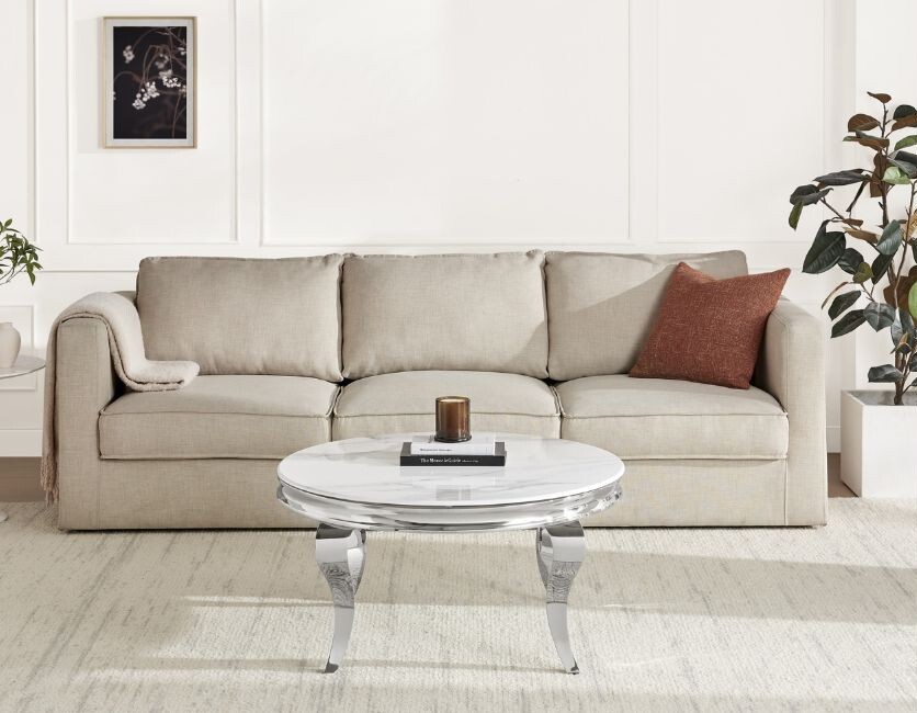 silver and white marble round coffee table with curved legs in front of beige fabric sofa with orange cushions, and orange glass cnagle holder on tabletop
