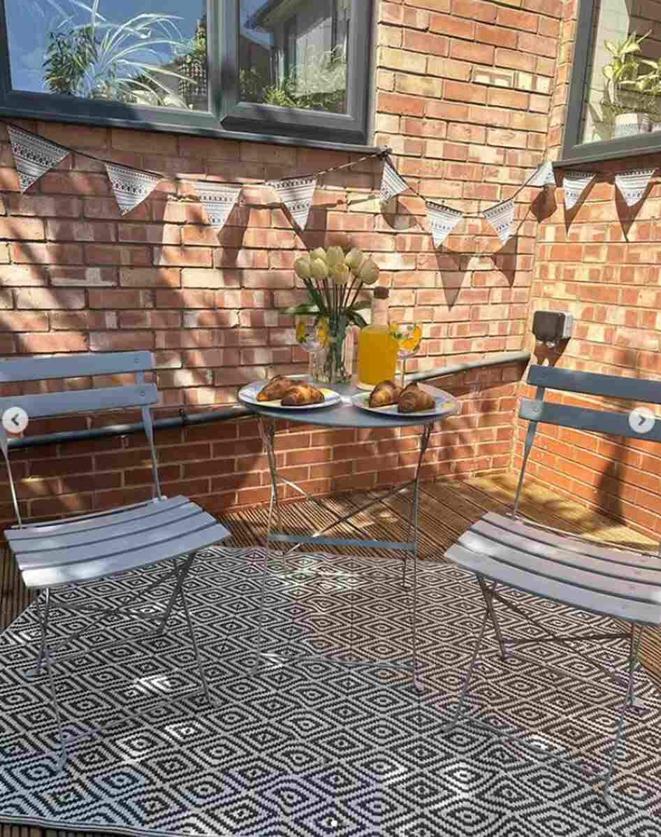 2 seat grey metal garden bistro seat on outdoor rug with brick wall and bunting behind and orange juice on table