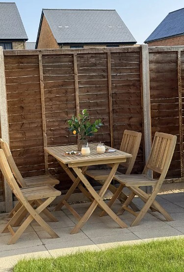 rectangular wooden slatted garden table with 4 matching wooden chairs on paving slab beside lawn and fence, with house roofs behind