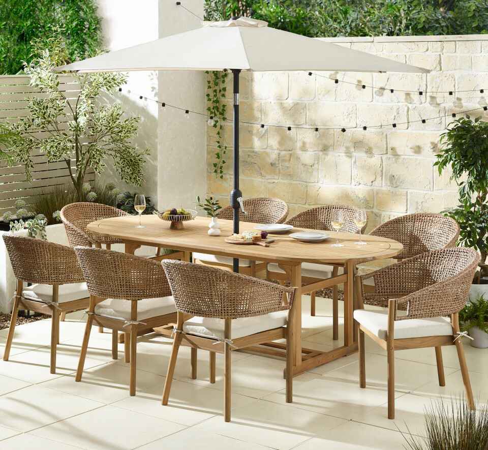 wooden oval garden table 8 wood and wicker chairs, paved patio, parasol, planting and fairy lights behind
